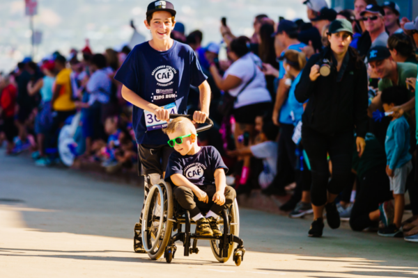 Young athlete with a physical disability, Brantley Volpel, rolls forward in his wheelchair during a Challenged Athletes Foundation Kids Run event as spectators line the course.