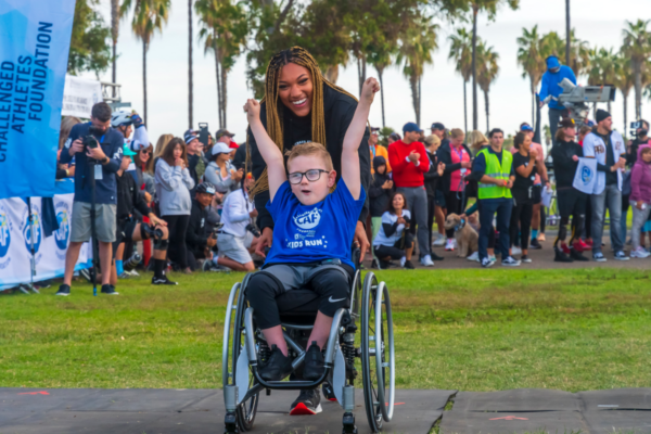 Brantley Volpel celebrates in his wheelchair during a Challenged Athletes Foundation Kids Run event as supporters, volunteers, and palm trees fill the background.