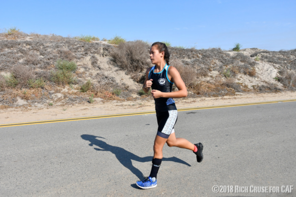 Athlete wearing a CAF triathlon kit runs along a paved road beside a sandy embankment under a clear sky.