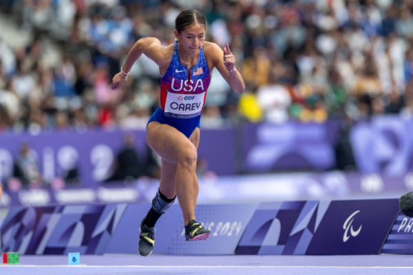 Track athlete wearing a USA uniform and a prosthetic lower leg runs on a track during a competition with blurred spectators in the background.
