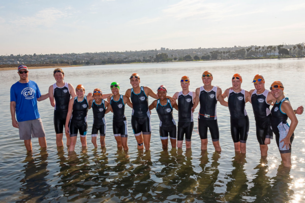 Group of athletes and a coach stand in shallow water wearing matching triathlon suits and swim caps, with a shoreline and buildings visible across the water.