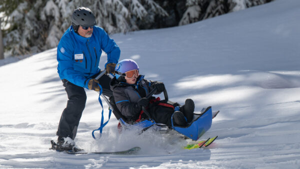 A man guiding a child in a blue adaptive ski chair down a snowy slope.