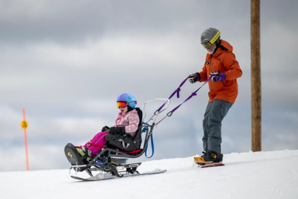 Adult guiding a child in a specialized ski chair down a snowy slope.