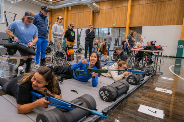 People participating in an indoor shooting training event in a gymnasium, with some lying prone and aiming rifles, while others observe.