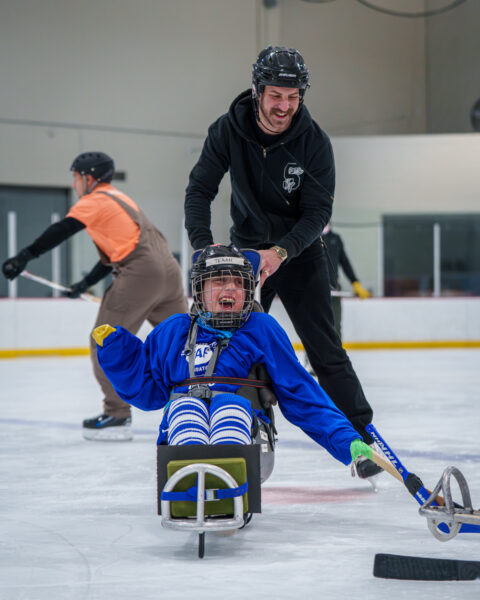 Person in a sled on ice being pushed by another individual during sled hockey practice.