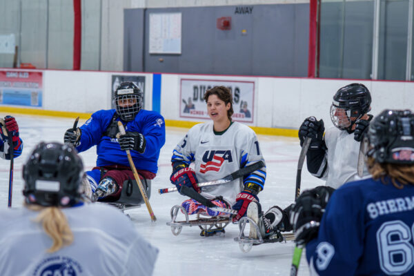 Athletes in sleds during a sled hockey session on an ice rink, with one wearing a "USA" jersey.