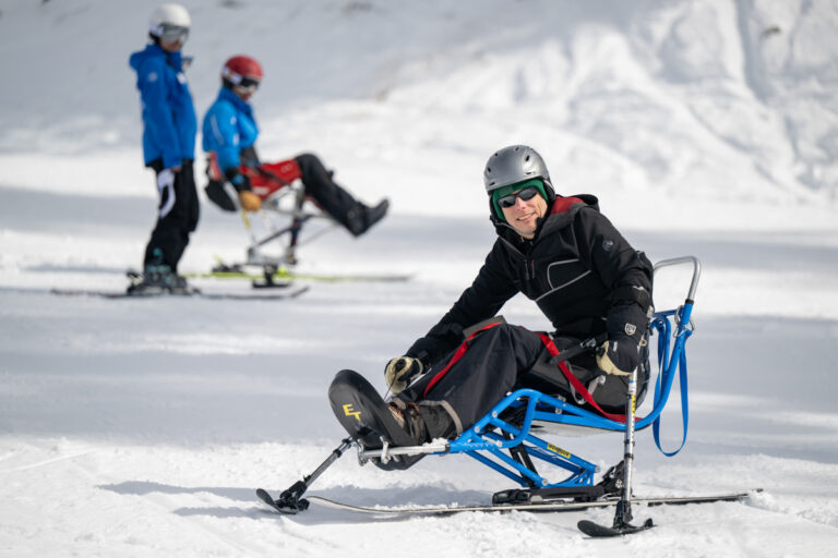 People engaged in adaptive skiing on a snowy slope, including one person in a blue sled in the foreground.