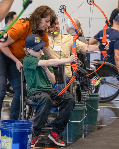 A young boy in a wheelchair aims a compound bow during an archery session, assisted by an adult.