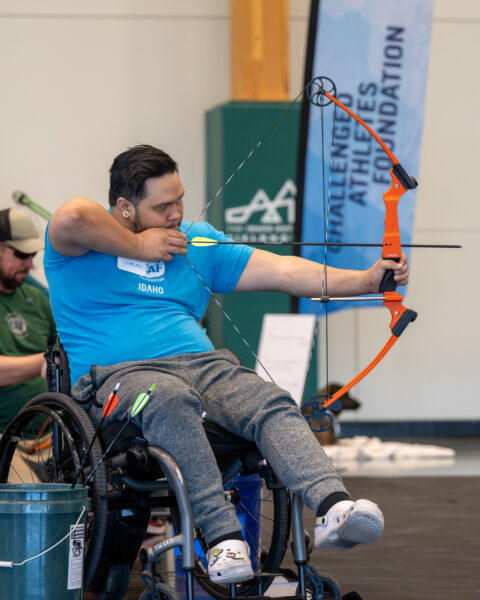 A person at an archery clinic in a wheelchair, aiming a bow and arrow.