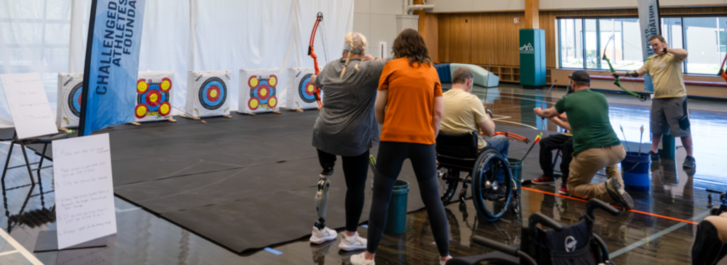 Adaptive archery session in a gym with participants, including a woman with a prosthetic leg and a man in a wheelchair, aiming at targets.