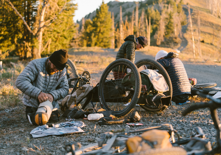 Athletes with physical disabilities preparing gear beside adaptive bicycles at a campsite, surrounded by forest and mountains in early morning light.