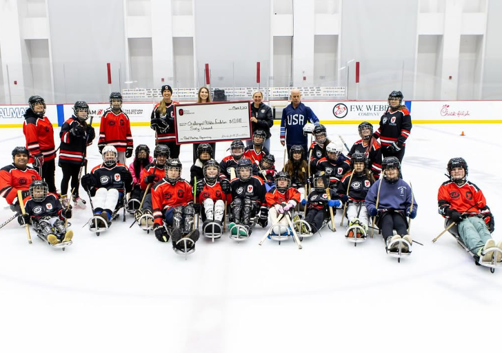 Group of youth athletes with physical disabilities participating in a sled hockey clinic, posing on the ice with coaches and partners while holding a $60,000 donation check at an indoor rink.