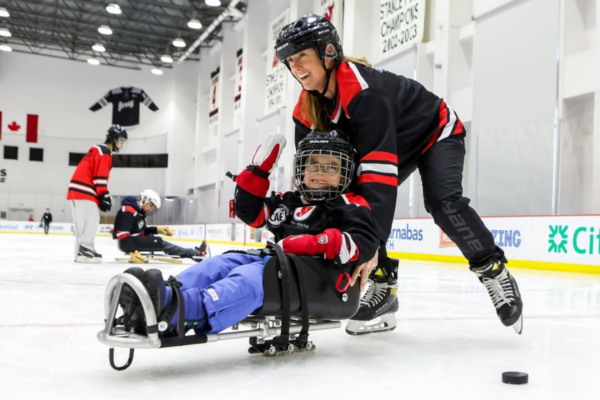 Coach skating behind a young athlete with a physical disability in a sled hockey sled, guiding them across the ice during a clinic session.