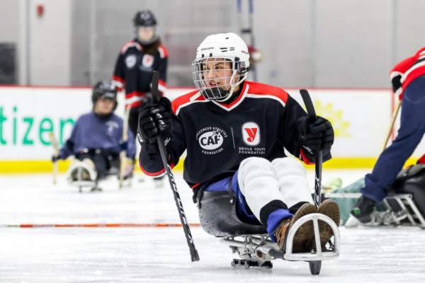 Youth athlete with a physical disability propelling a sled hockey sled forward using sticks on the ice during a skills drill.