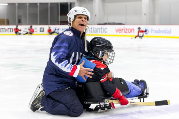Coach in a USA hockey jersey kneeling on the ice while assisting a young athlete with a physical disability in a sled hockey sled.