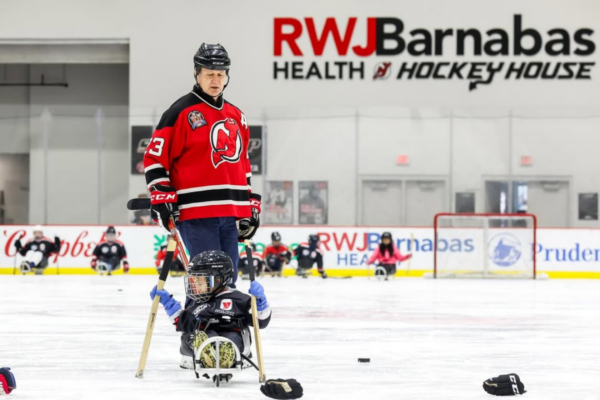 Coach in a USA hockey jersey kneeling on the ice while assisting a young athlete with a physical disability in a sled hockey sled.