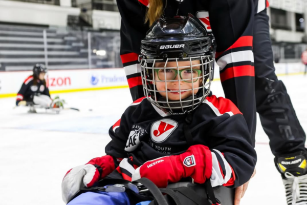 Close-up of a smiling youth athlete with a physical disability wearing a red sled hockey jersey and holding sticks while seated in a sled on the ice.