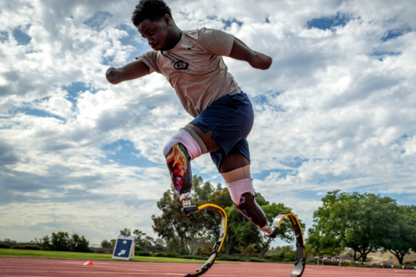 A young athlete with a physical disability runs on a track using running blades, captured mid-stride against a bright sky.
