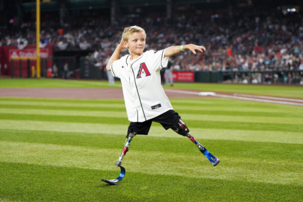 A young athlete with a physical disability throws a baseball on a professional field, wearing a team jersey during a stadium event.