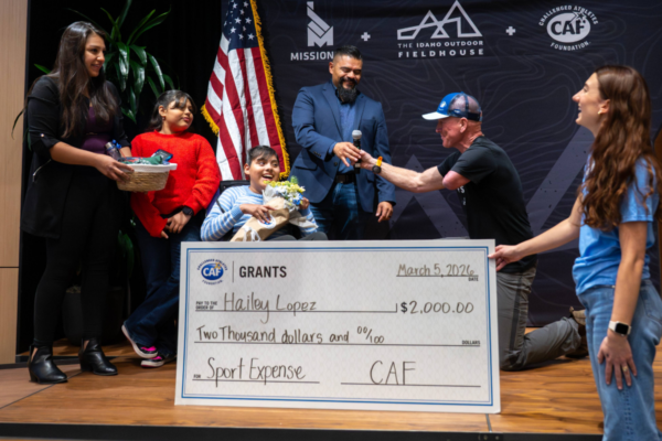 A CAF grant recipient with a physical disability sits on stage holding a large check, surrounded by supporters during a grant presentation ceremony.