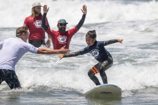 A young athlete with a physical disability rides a wave while instructors cheer and guide them in the ocean.