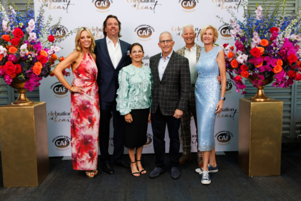 A group of guests pose together in front of a Celebration of Heart step-and-repeat backdrop framed by large floral arrangements.