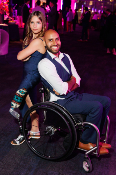 Two gala attendees pose back-to-back on the event floor, one seated in a wheelchair and the other standing, with pink lighting and other guests mingling in the background.