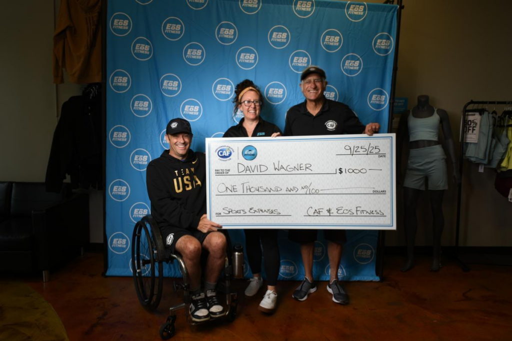 Grant recipient in a wheelchair holding a large check with supporters indoors.