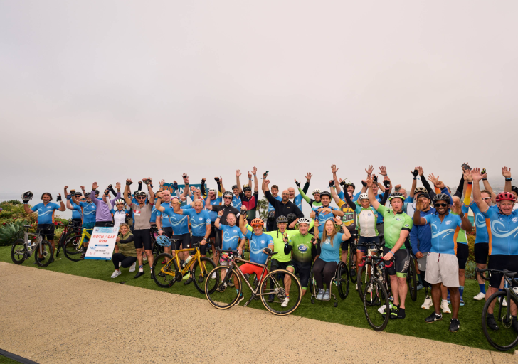 Large group of CAF cyclists and participants standing with bicycles on a grassy overlook, many raising their arms, with a coastal landscape visible in the background.