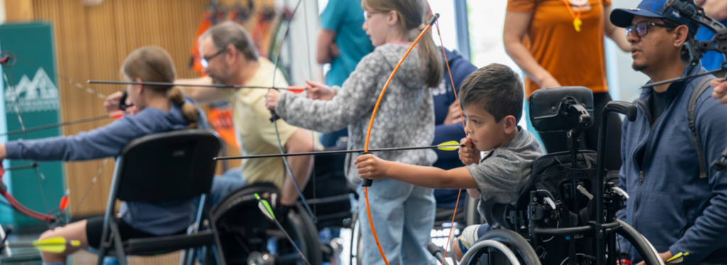 Group of people, including a young boy in a wheelchair, practicing archery indoors.
