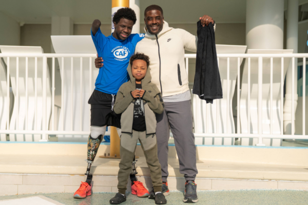 Three people standing together near a poolside railing, one with a prosthetic leg wearing a blue CAF shirt, another holding a towel, and a child standing in front, all facing the camera.