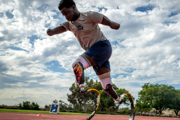Athlete with prosthetic running blades mid-jump on a track, wearing a gray shirt and blue shorts, with a cloudy sky and trees in the background.