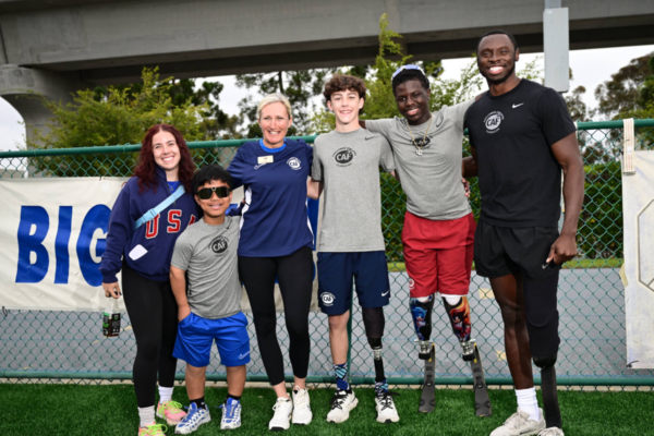 Group of six people standing together in front of a fence outdoors, some wearing CAF apparel, including individuals with prosthetic legs, all facing the camera.
