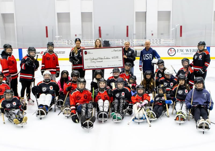 Group of youth sled hockey players and adults posing on an indoor ice rink holding a large presentation check with rink boards and sponsor signage in the background