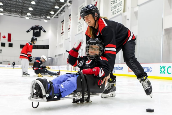 Adult pushing a young sled hockey athlete seated in a sled across the ice while other players practice in the background