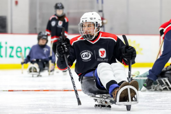 Close view of a sled hockey athlete in a Jersey holding two sticks while moving across the ice with other players behind