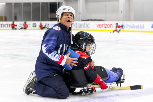 Adult kneeling on ice behind a young sled hockey athlete seated in a sled and holding a hockey stick during practice