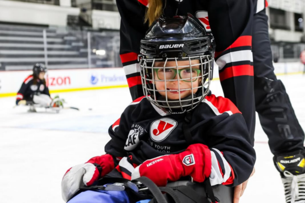 Close-up of a young sled hockey athlete seated in a sled wearing helmet and gloves with another person positioned behind on the ice