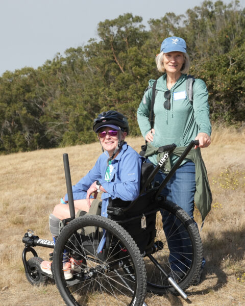 Two people outdoors, one seated in an off-road wheelchair, smiling, with trees in the background.