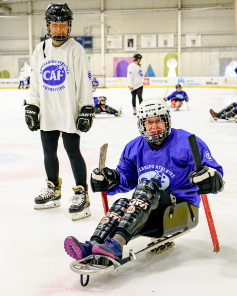 Adaptive hockey athletes practicing on ice; one sits in a sled while another stands with skates, both in jerseys.