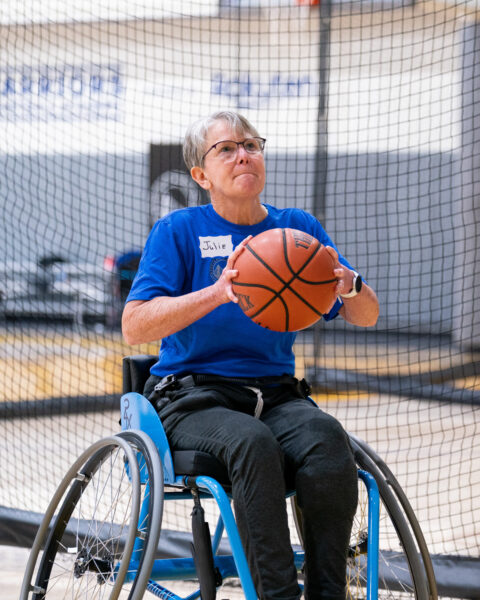 An adult in a sports wheelchair holds a basketball in a gym.