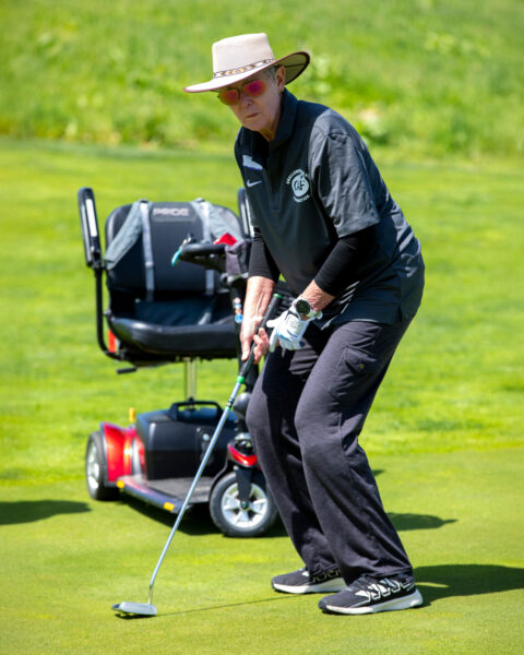 Person preparing to putt on a golf course with a mobility scooter nearby.