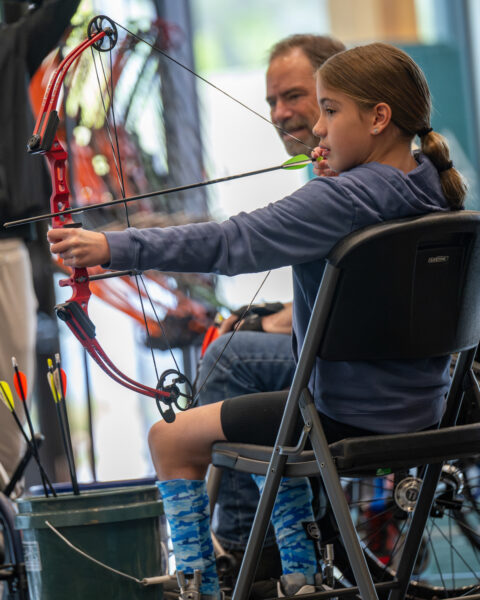 A young person aims a red compound bow indoors, with an adult supervising nearby.