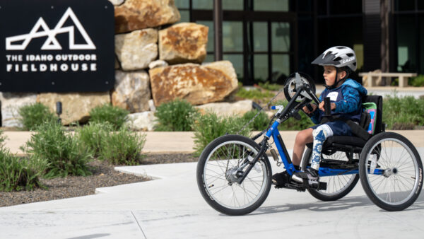 Child on a blue adaptive handcycle near a sign that reads "The Idaho Outdoor Fieldhouse."