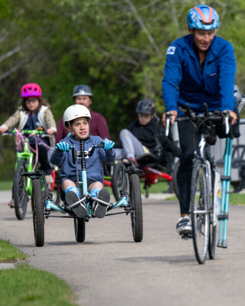 Group of people riding bicycles and tricycles on a path in a park area.