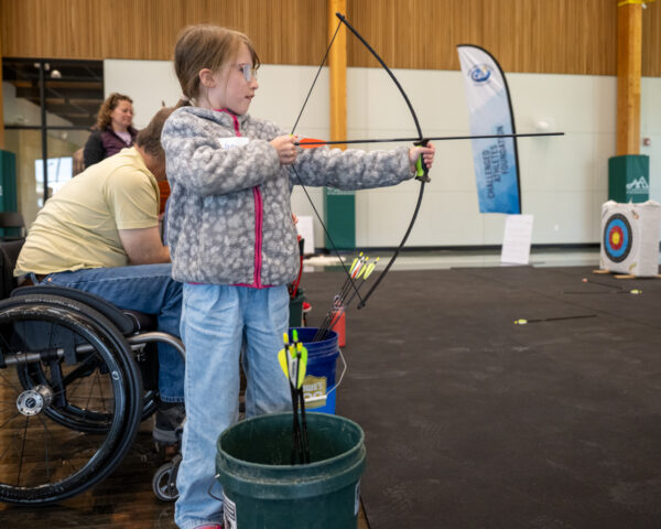 Young girl practicing archery indoors near a person in a wheelchair and an archery target.