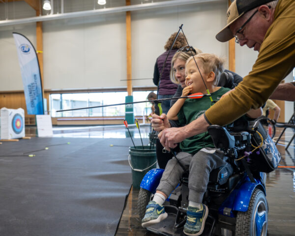 Child in wheelchair practicing archery with adult assistance in a large indoor space.
