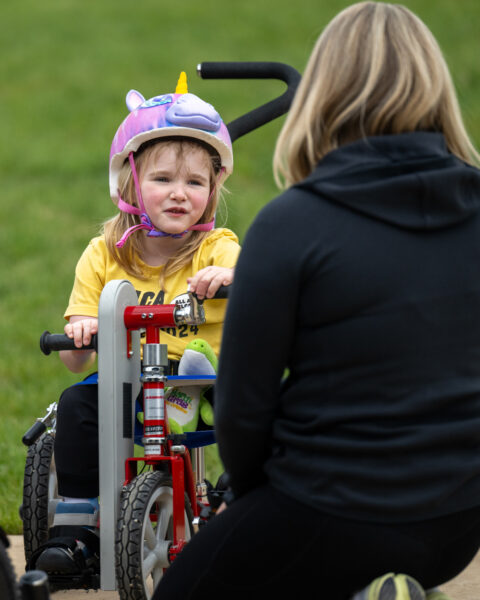 Young girl on a red adaptive tricycle, wearing a unicorn helmet, interacting with an adult woman.