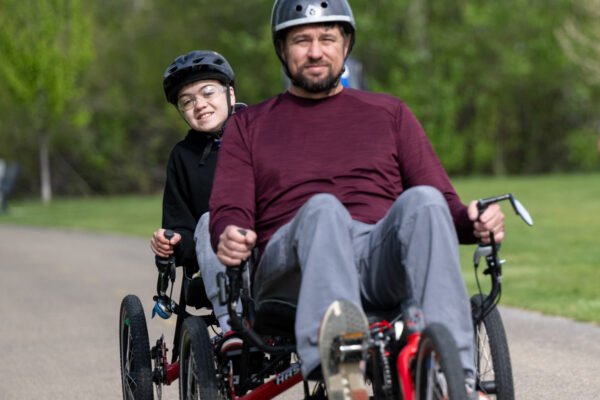 Two people riding red recumbent bikes on a paved path in a park, wearing helmets.