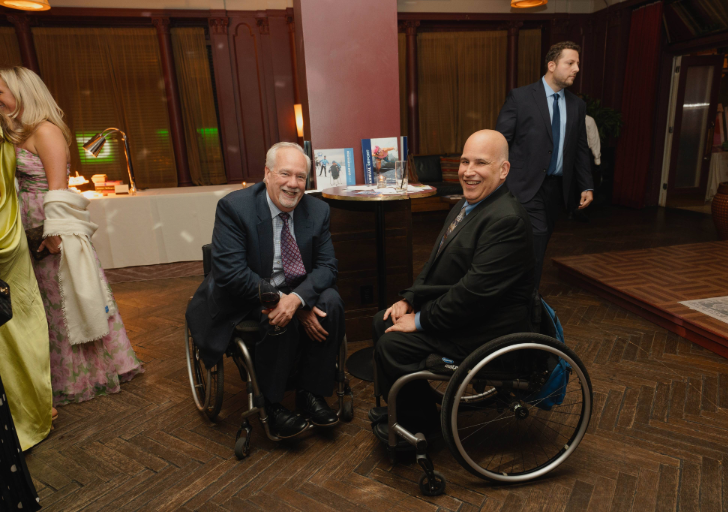 Two attendees in suits seated in wheelchairs at a cocktail table with CAF materials displayed in the background